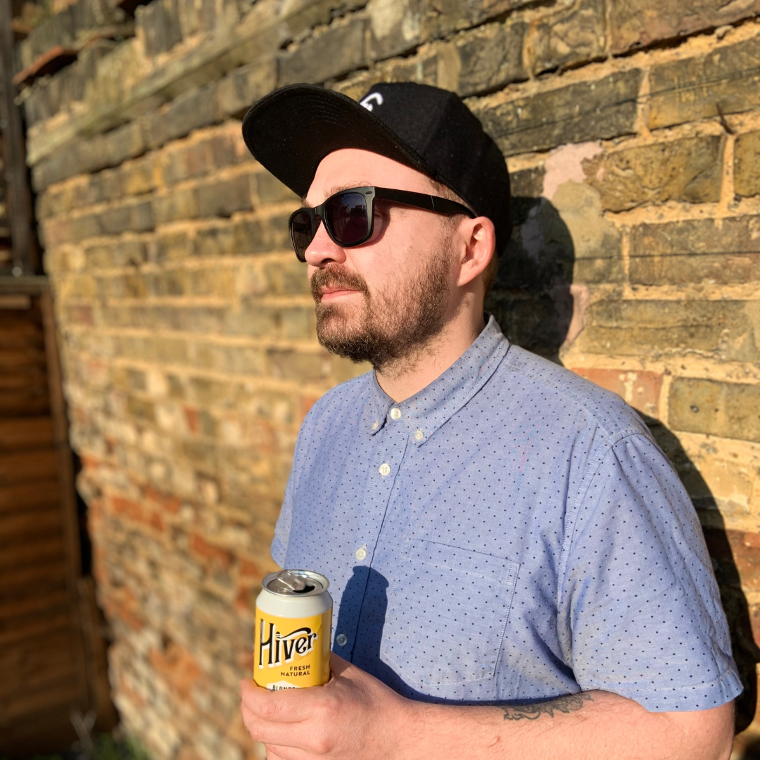 Man wearing black Phoebee cap and sunglasses holding a can of Hiver beer against brick wall