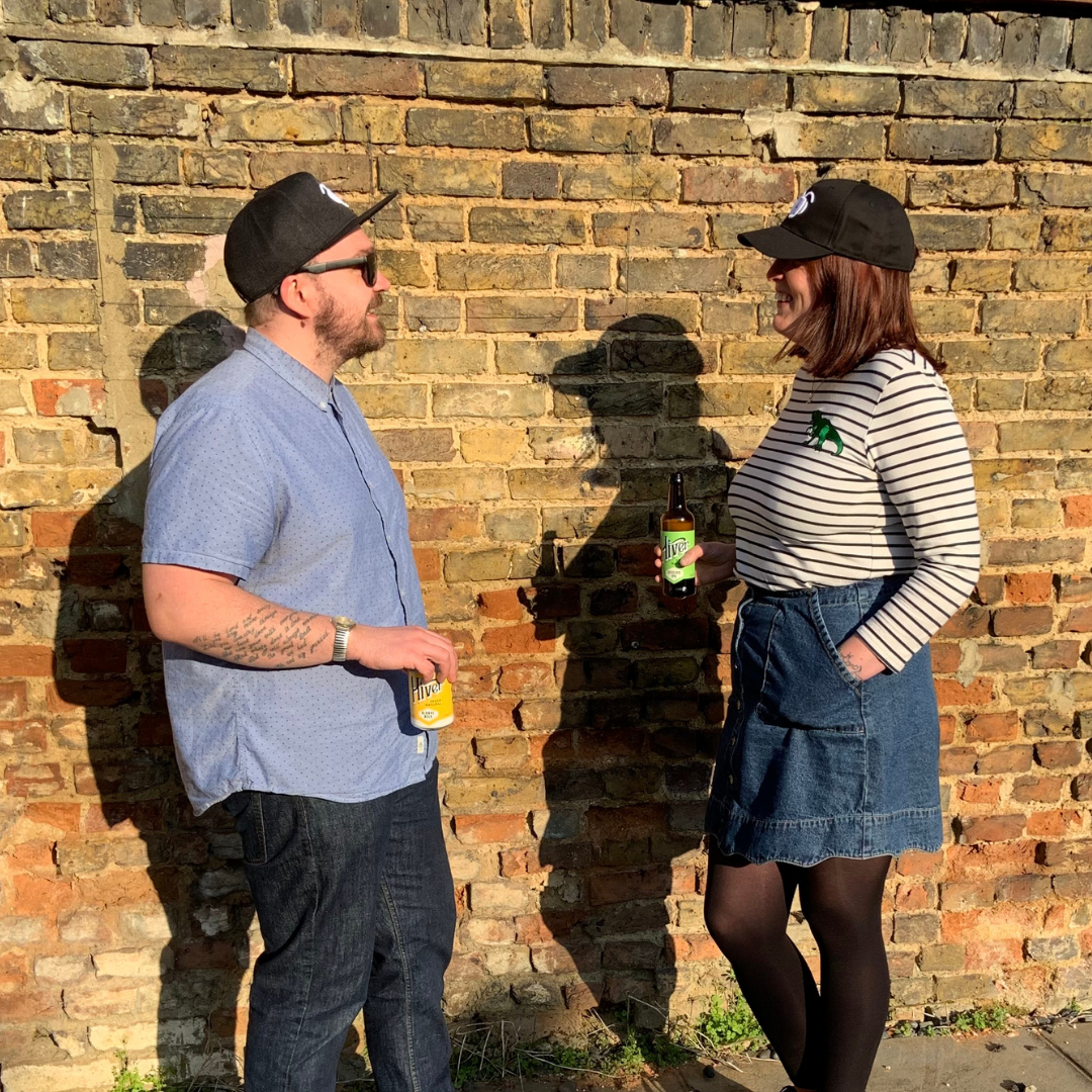 Man and woman wearing black Phoebee caps chatting and holding Hiver drinks against brick wall
