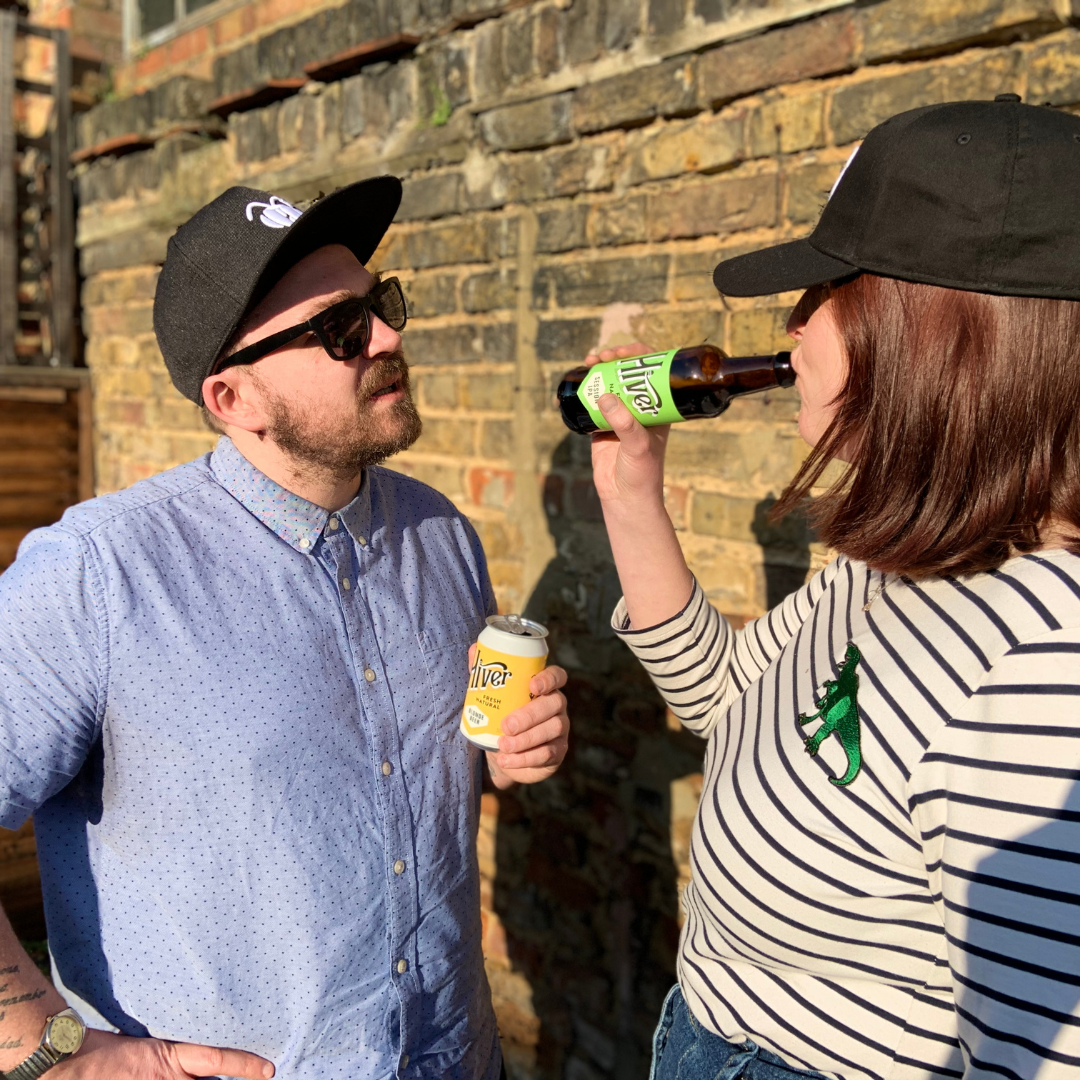 Man and woman wearing black Phoebee caps drinking Hiver can and bottle against a brick wall