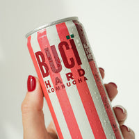 Hand holding a can of Buci Hard Kombucha with red and white striped design.