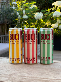 Three cans of BUCI Hard Kombucha on a table in a floral outdoor setting.