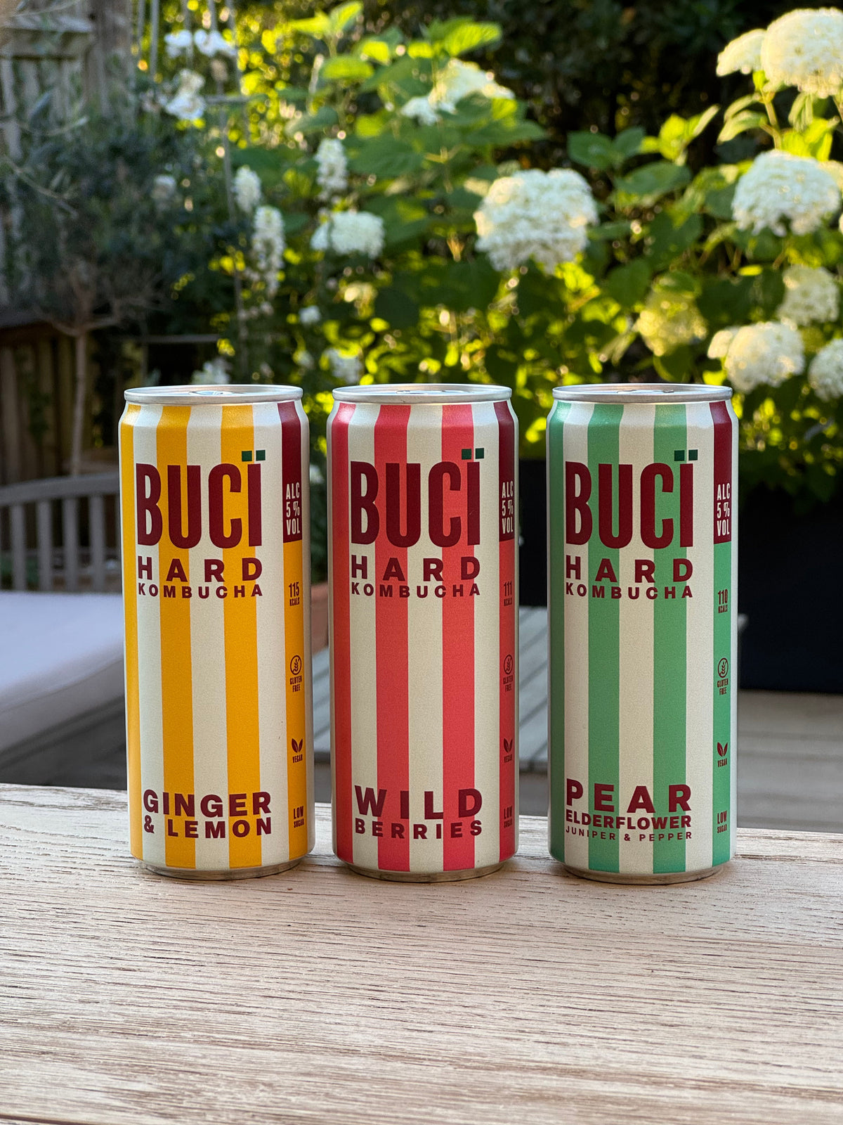 Three cans of BUCI Hard Kombucha on a table in a floral outdoor setting.