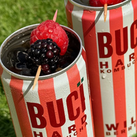 Two cans of BUCÏ Wild Berries Hard Kombucha with berries on a grassy background.