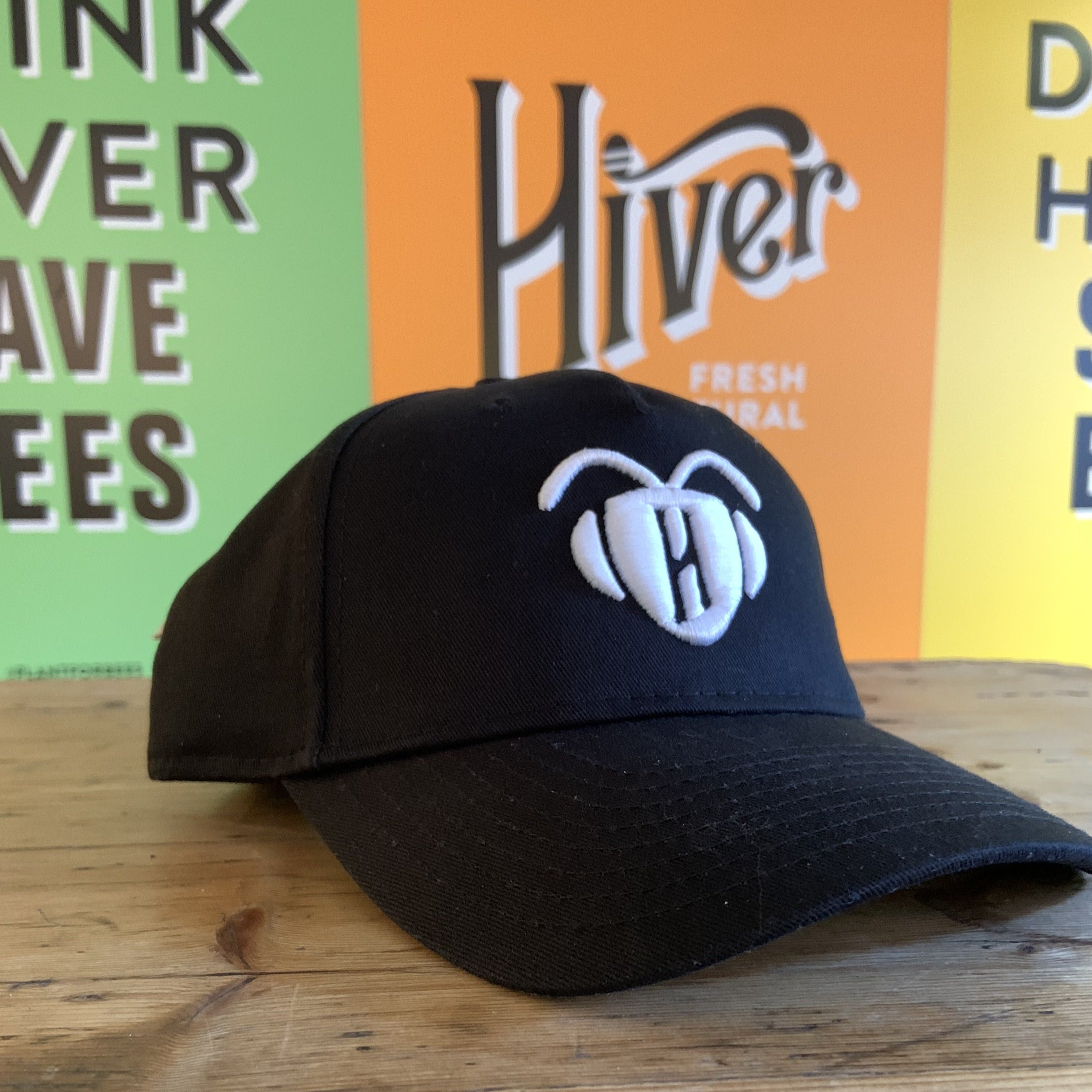 Black Phoebee cap with white embroidered bee logo on wooden table in front of Hiver branding
