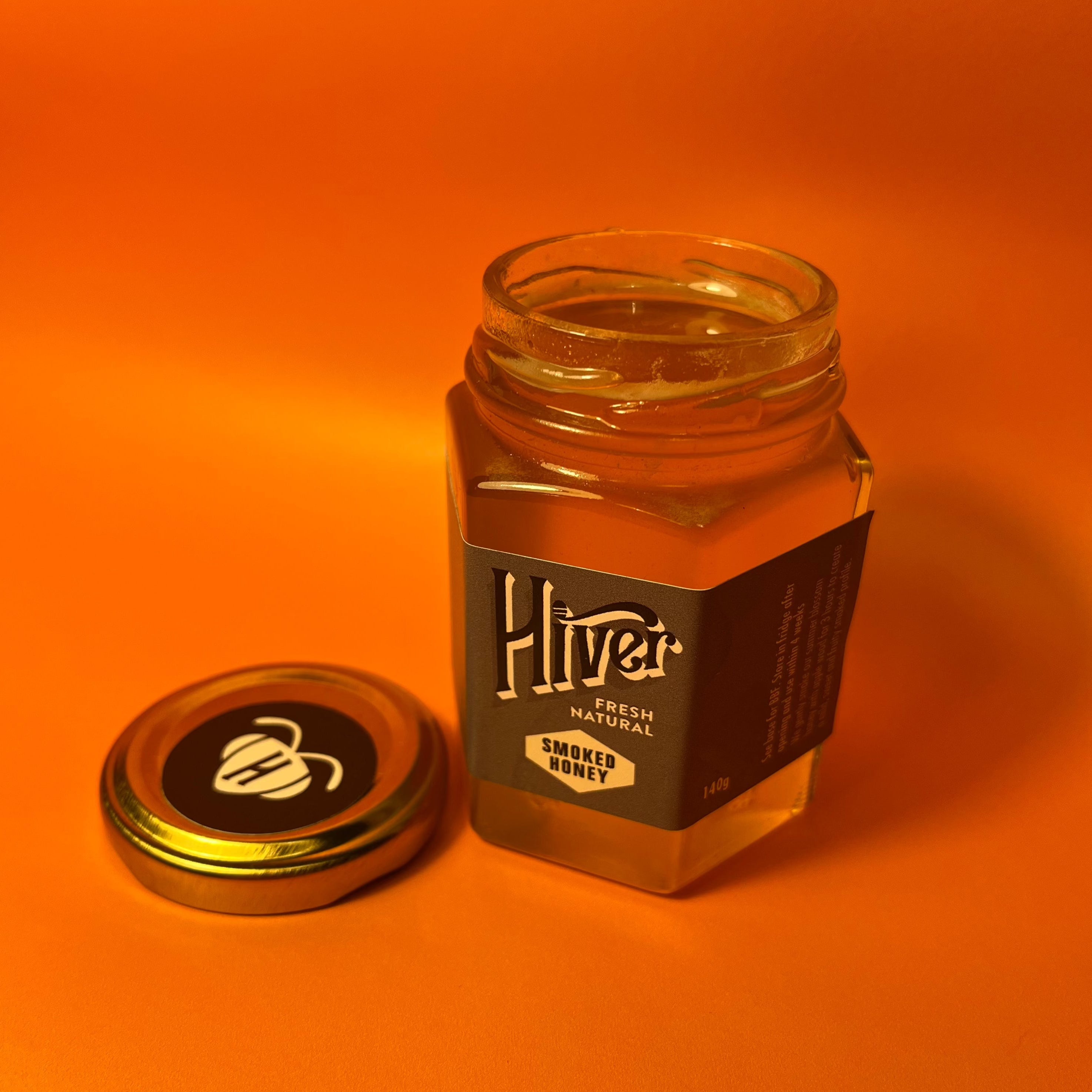 Open jar of Hiver Smoked Honey with lid removed beside it on an orange background